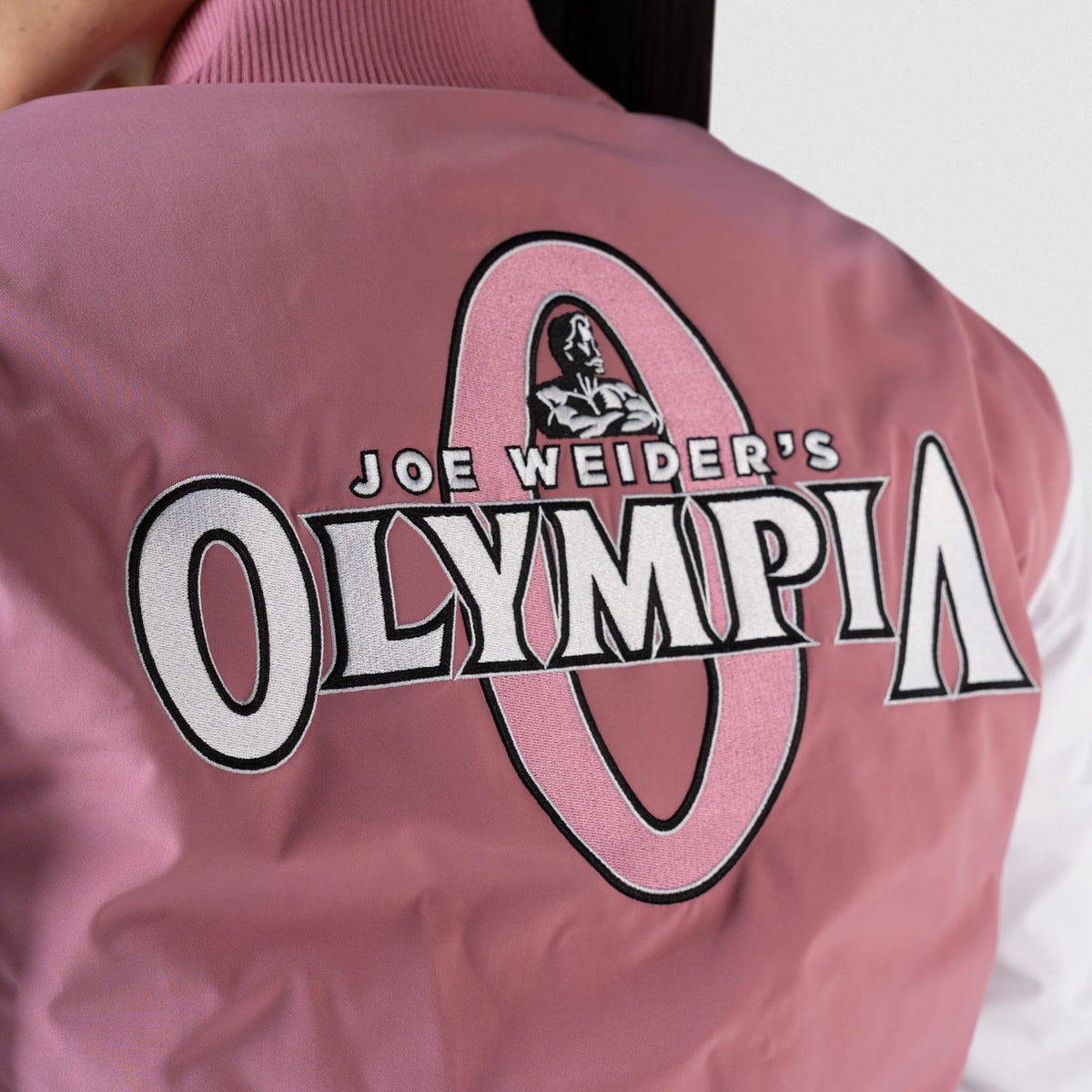 Rose Division Crop Bomber Jacket | Back logo detail of the Joe Weider's "Olympia" logo in white stitching on the pink jacket