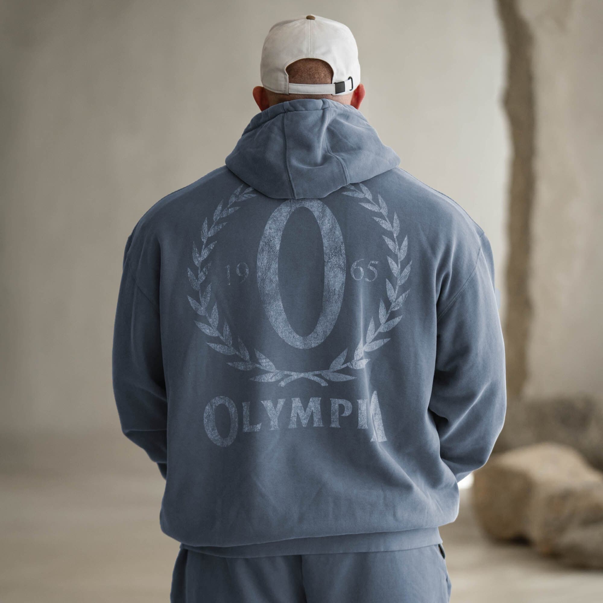 Model wearing pale blue Olympia hoodie, back view highlighting the oversized wreath "O" graphic in white.