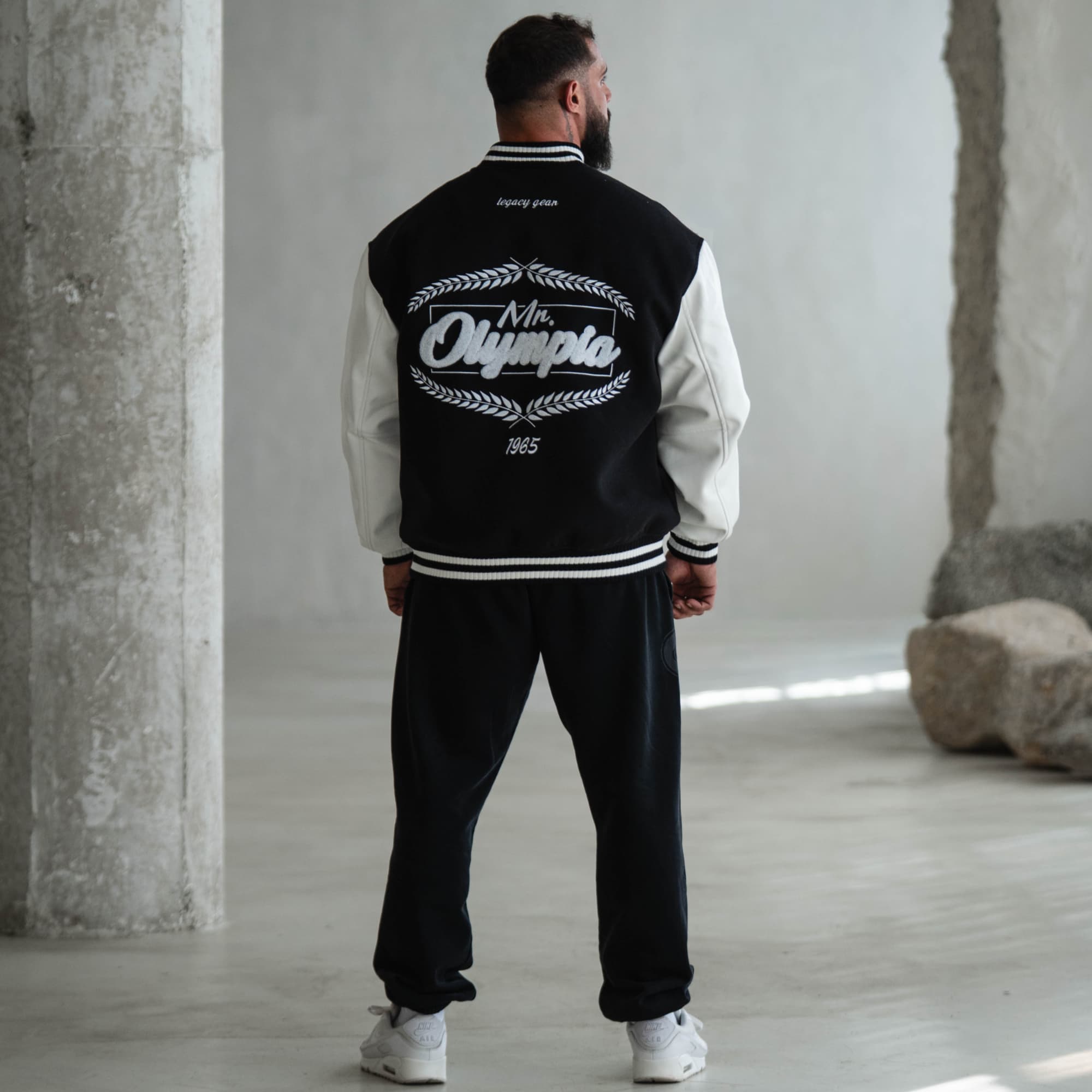 Model wearing Mr. O Varsity Jacket, back view showing oversized embroidered “Mr. Olympia 1965” graphic.