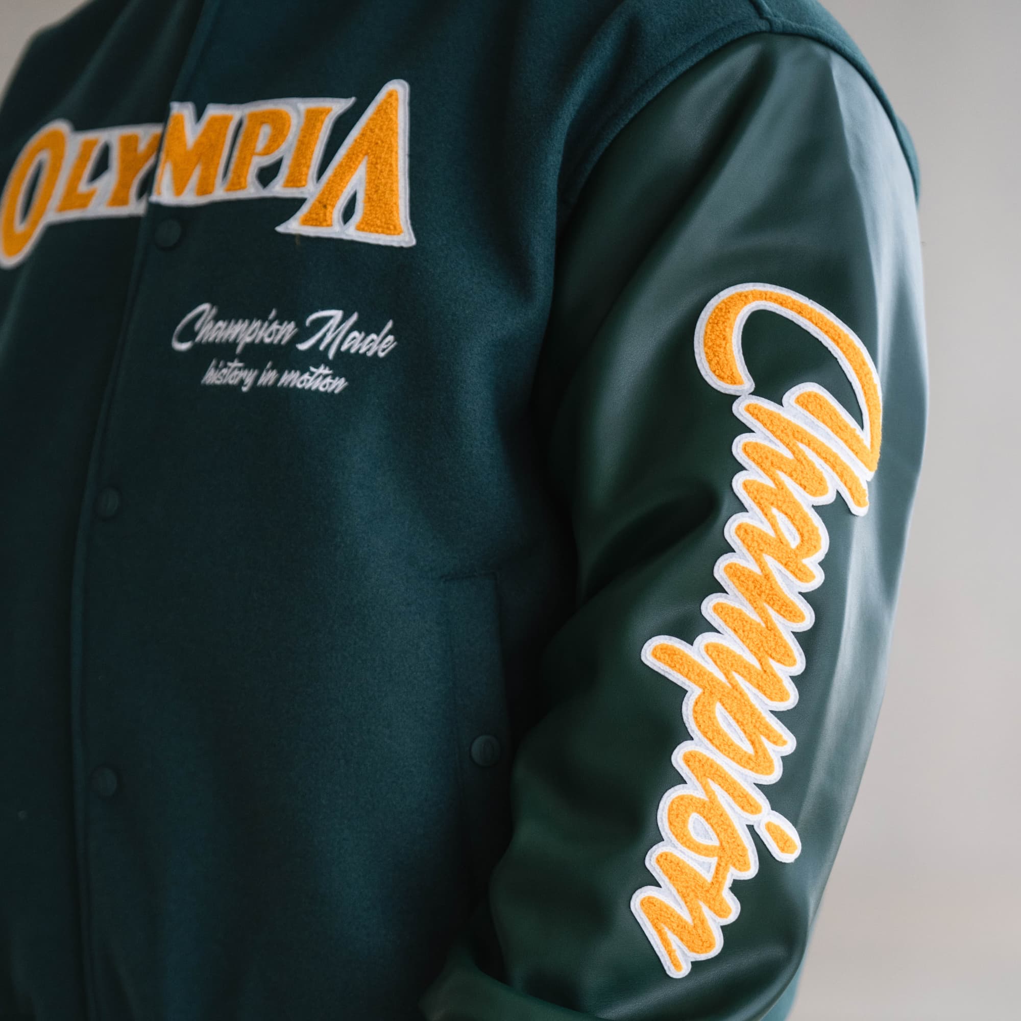 Side view close-up of embroidered “Champion” text on the sleeve of the green Olympia Champion Made Varsity Jacket.