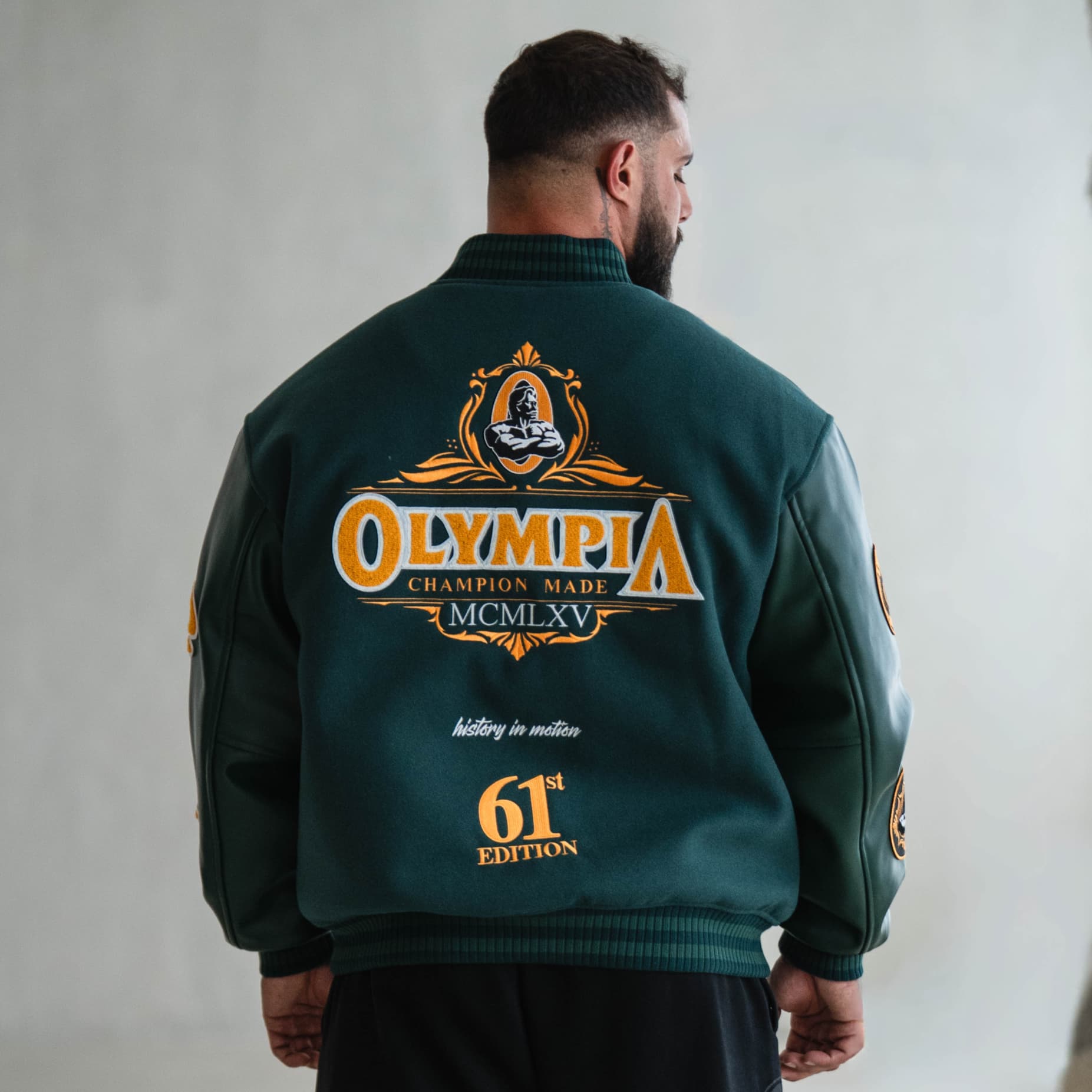 Back view of green Olympia Champion Made Varsity Jacket with bold gold embroidery reading “Olympia Champion Made MCMLXV” and “61st Edition.”