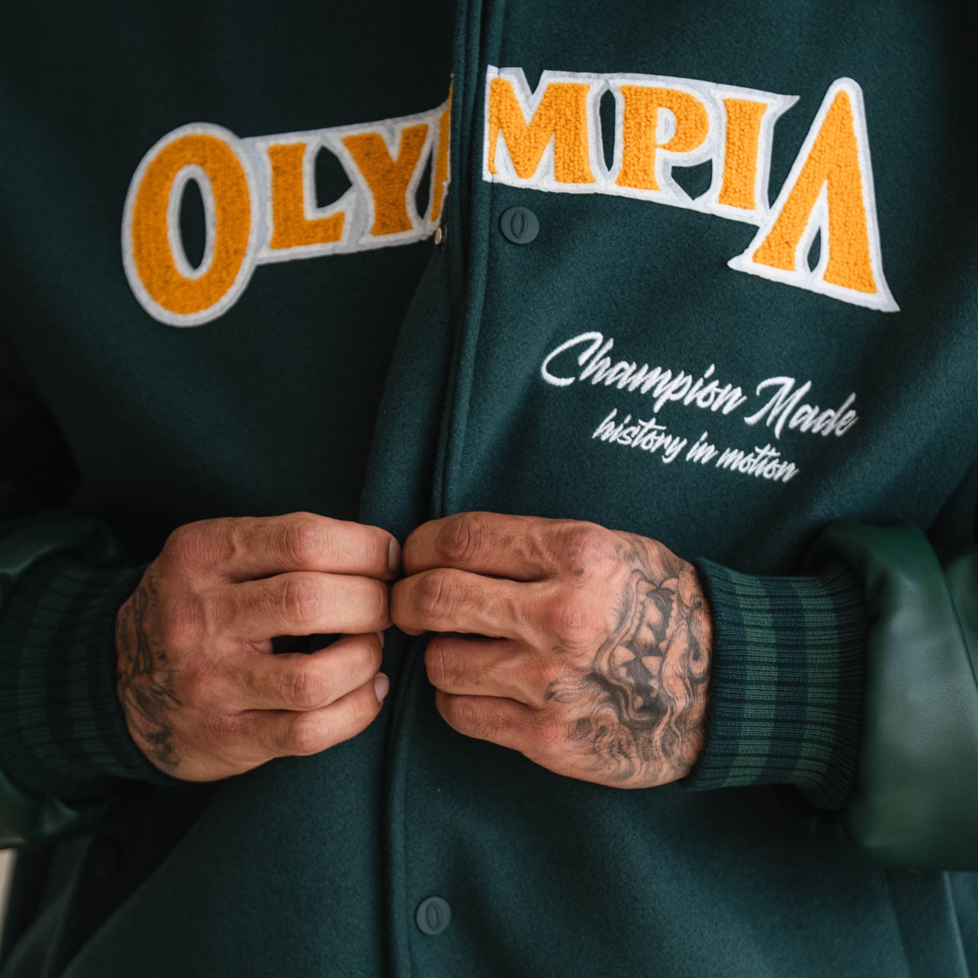 Close-up of hands fastening the Olympia Champion Made Varsity Jacket, highlighting chest embroidery details.