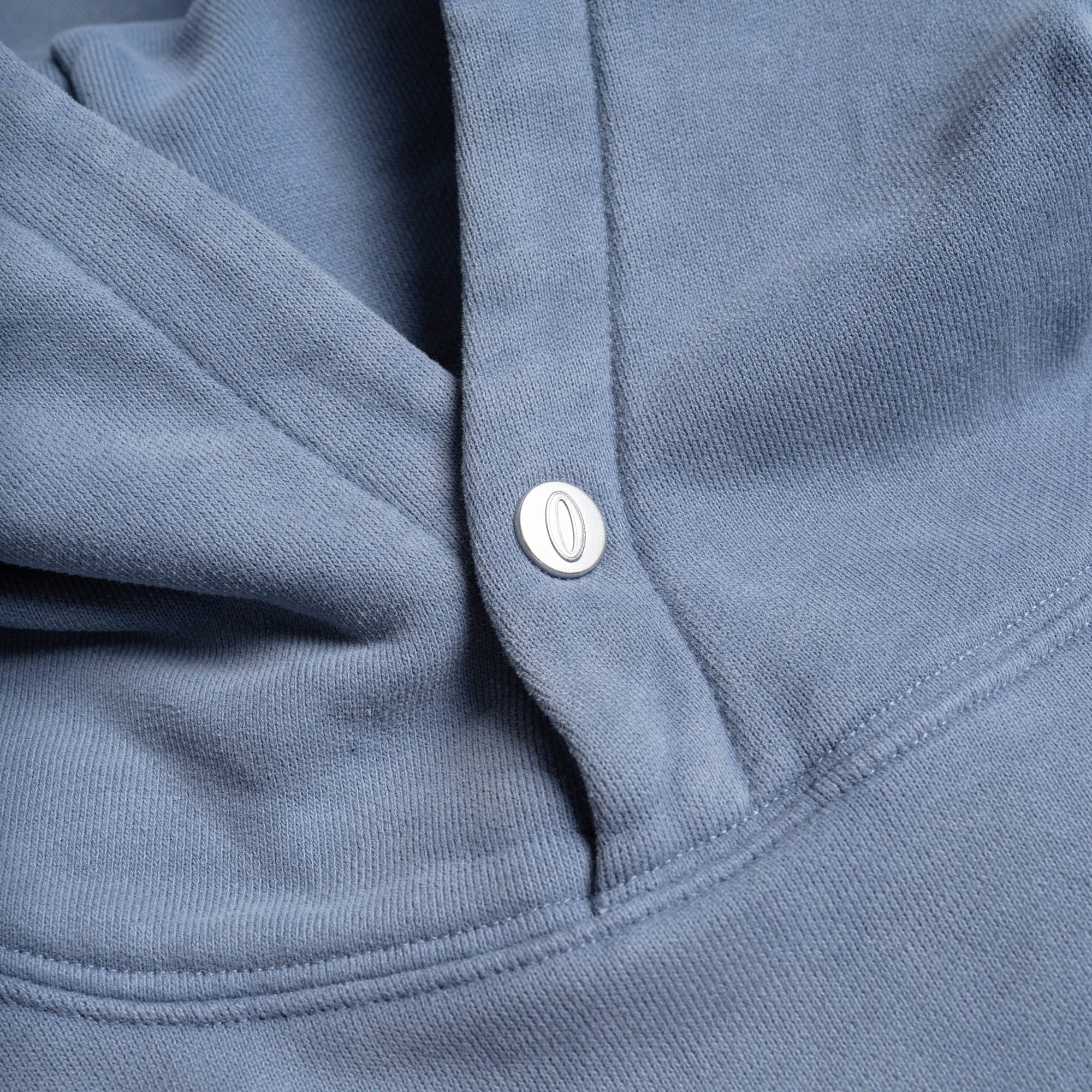 Detail shot of the pale blue hoodie hood with branded snap button closure featuring embossed "O" logo.